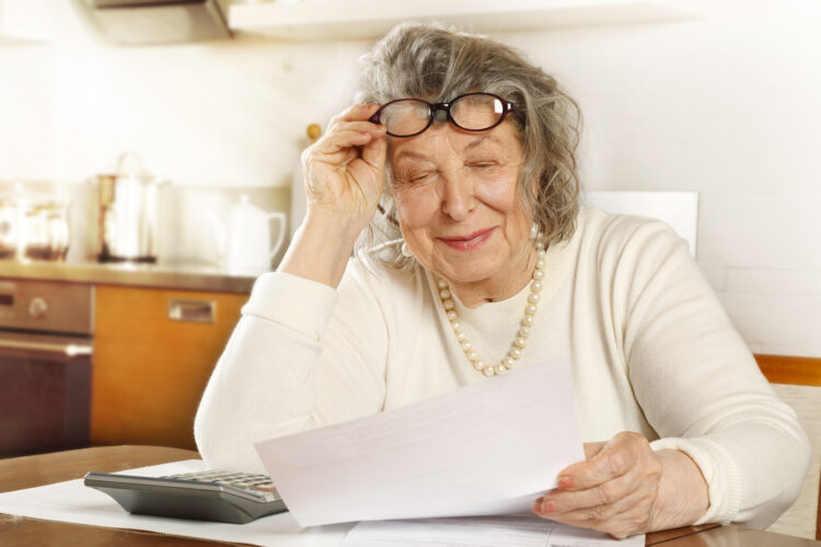 An old lady sitting at a table in the kitchen looking through receipts with a calculator