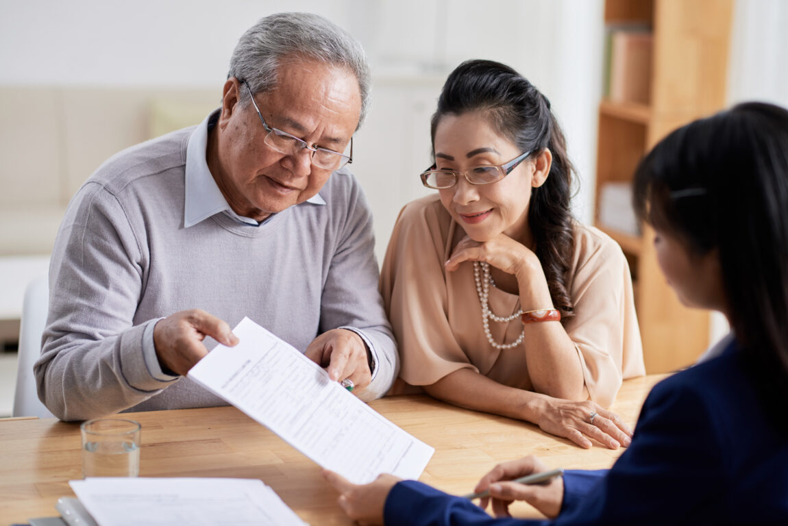 Concentrated senior man and his pretty wife studying terms of real estate purchase agreement while having meeting with Real Estate Agent, interior of modern office on background